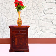 A brown solid wood bedside table with drawers, featuring a vase with red flowers on top, placed against a wall with a floral pattern.