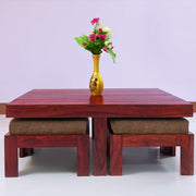 A rectangular solid wood coffee table with stool in brown color, placed on a white floor against a purple background, with a yellow vase on top of the table containing flowers.