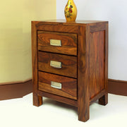 A wooden bedside table with three drawers, finished in provincial teak, with metal handles and a decorative vase on top.