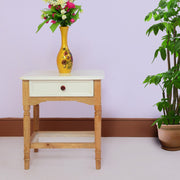 A wooden side end table with a drawer, finished in natural white, placed against a purple wall with a potted plant beside it. The table has a vase with flowers on top.