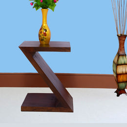 A wooden Z-shaped side end table in walnut finish placed on a white surface with a vase on top and decorative items in the background.