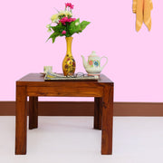 A wooden coffee table in a natural finish, with a vase of flowers and a teacup on top, placed against a pink wall.