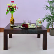 A square center coffee table made of walnut wood finish, with decorative items, a vase with flowers, and a tea set displayed on it. The table is placed against a purple backdrop with a potted plant to the right.
