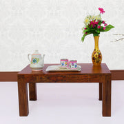 A solid wood center coffee table in a teak finish displayed with a vase of flowers, cups, and a teapot on top.