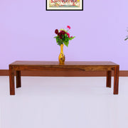 A wooden bench with a teak finish placed against a purple wall, with a vase of flowers on top.