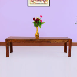 A wooden bench with a teak finish placed against a purple wall, with a vase of flowers on top.