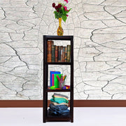 A wooden open bookshelf in a living room setting, with books, decorative items, and clothing items displayed on the shelves. The bookshelf has a dark finish, and there is a vase with flowers on top of it.