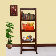 A wooden open bookshelf furniture with multiple items placed on its shelves, such as books, pillows, and decorative items. The bookshelf is positioned in a room with a plant and a framed picture on the wall in the background.
