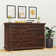 A wooden chest of drawers with multiple drawers, placed against a wall with decorative framed pictures, next to a potted plant. The chest has a rich wood finish and metal handles on the drawers.