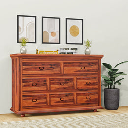 A wooden chest of drawers with multiple drawers, placed against a wall with decorative framed pictures, next to a potted plant. The chest has a rich wood finish and metal handles on the drawers.