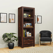 A wooden bookshelf with multiple shelves and a drawer, placed in a room with a chair and a potted plant, displaying a variety of books.