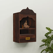 A wooden wall-mounted temple with a drawer, featuring a sculpture of a deity on the shelf, set against a white wall with a green plant beside it.