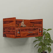 A wooden wall-mounted temple with a drawer, featuring a Buddha statue and LED lights, mounted on a wall beside a green plant.