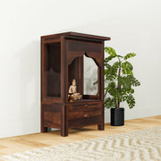 A wooden temple with a drawer, crafted in Sheesham wood with a mirror on the door, placed on a floor with a plant beside it.