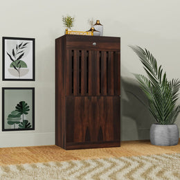 A wooden shoe rack with a drawer, finished in dark brown, placed against a wall with decorative items on top, situated in a bright room with a potted plant to the right.