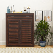 A brown solid wood shoe rack with a drawer, placed in a living room setting with decorative items on top and a potted plant beside it.